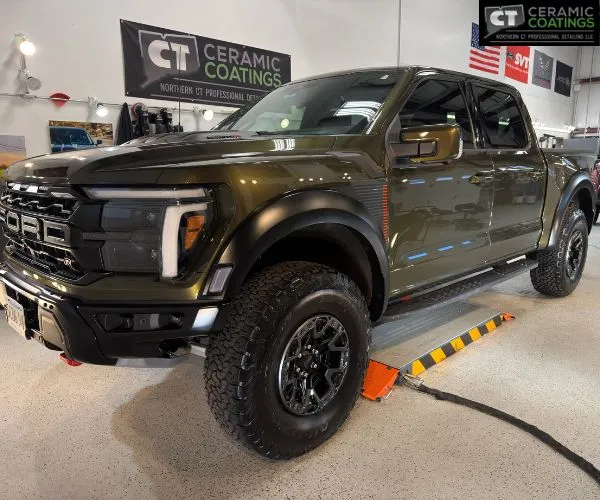 Front three-quarter view of a olive-green Ford F-150 Raptor pickup truck inside a auto detailing garage.