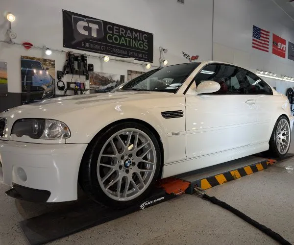 Front side view of a glossy white BMW M3 coupe with silver mesh wheels inside a detailing shop on a lift ramp.